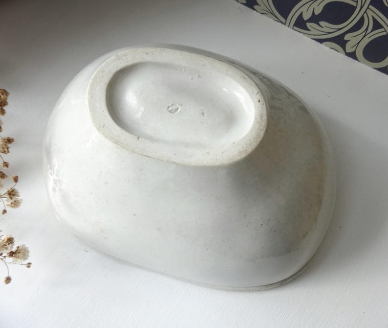May include: A white, oval-shaped ceramic bowl with a slightly textured surface. The bowl has a wide rim and a smooth interior. The underside of the bowl is visible, showing the base and a small marking. The bowl is likely vintage.