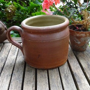 May include: A brown ceramic crock with a handle and a light brown glaze on the rim. The crock is sitting on a wooden table.
