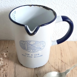 Pode incluir: Jarro de esmalte branco com alça e borda azul escuro. O jarro tem um bico e um gráfico azul e branco com o texto "COW & GATE MILK FOOD MEASURE". Uma colher de prata está em uma superfície de madeira.