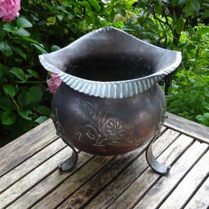 Pode incluir: Um vaso de cobre marrom escuro com um design floral decorativo e três pernas. O vaso está sobre uma superfície de madeira.