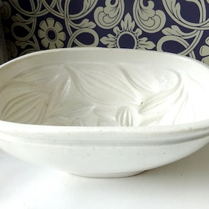 May include: A white ceramic oval bowl with a textured floral design. The bowl has a wide rim and a slightly speckled surface. The background includes a blue and white patterned wallpaper and a decorative ceramic vase.