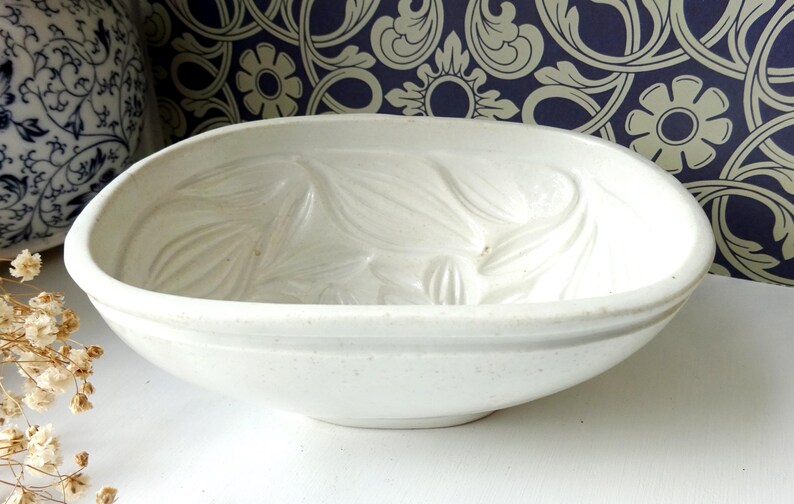 May include: A white ceramic oval bowl with a textured floral design. The bowl has a wide rim and a slightly speckled surface. The background includes a blue and white patterned wallpaper and a decorative ceramic vase.