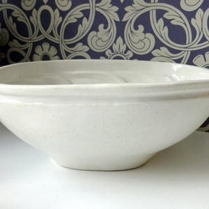 May include: A speckled, off-white ceramic bowl with a rounded rectangular shape. The bowl has a slightly flared rim and sits on a white surface. The background features a blue and cream floral patterned wallpaper.