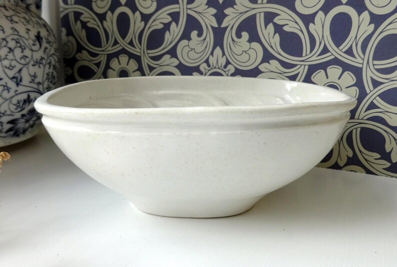 May include: A speckled, off-white ceramic bowl with a rounded rectangular shape. The bowl has a slightly flared rim and sits on a white surface. The background features a blue and cream floral patterned wallpaper.