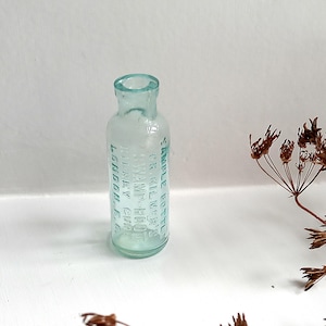 Puede incluir: Una pequeña botella de vidrio azul claro antigua con texto en relieve. La botella tiene forma cilíndrica con cuello estrecho y borde ligeramente ensanchado. El texto parece ser un nombre de producto y ubicación. Plantas secas en el fondo.