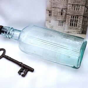 May include: A clear glass bottle with a textured surface and a brown cap. The bottle is lying on a white surface next to a rusty metal key.