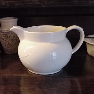 以下が含まれることがあります： 丸みを帯びたボディとハンドルが付いた白いセラミックピッチャー。ピッチャーは、他のセラミック容器のある木製の表面の上に置かれています。