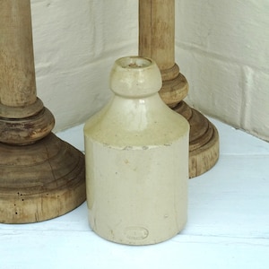 May include: A cream-colored, antique stoneware bottle with a rounded shoulder and cylindrical body. The bottle is displayed with two wooden candlestick holders. The background is a white painted brick wall and a white surface.