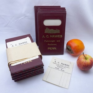 May include: A stack of burgundy-colored paper notebooks with gold lettering that reads "A. C. HAWES Fishmonger and Fruiterer, PENN."  A stack of similar notebooks is wrapped in a cream-colored paper band.  A persimmon and a red apple are on a white surface.