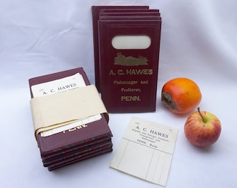 May include: A stack of burgundy-colored paper notebooks with gold lettering that reads "A. C. HAWES Fishmonger and Fruiterer, PENN."  A stack of similar notebooks is wrapped in a cream-colored paper band.  A persimmon and a red apple are on a white surface.