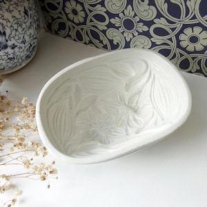 May include: A white ceramic dish with a floral design. The dish has a curved, oval shape and is decorated with embossed leaf-like patterns. The background includes a blue and white patterned wallpaper and a decorative ceramic vase.
