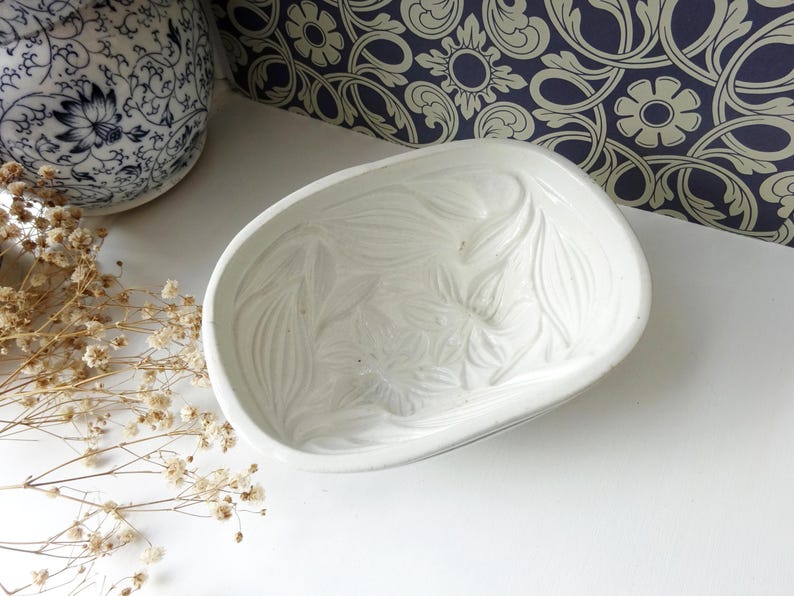 May include: A white ceramic dish with a floral design. The dish has a curved, oval shape and is decorated with embossed leaf-like patterns. The background includes a blue and white patterned wallpaper and a decorative ceramic vase.