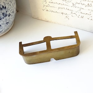 Può includere: Un oggetto decorativo rettangolare in ottone con due spazi rettangolari aperti e un piccolo foro al centro. L'oggetto ha una parte superiore arrotondata e una parte inferiore leggermente curva.
