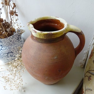 May include: A rustic, terracotta-colored pitcher with a rounded body and a handle. The rim is painted with a cream and brown glaze. The pitcher is likely vintage or antique, adding a touch of old-world charm to any decor.