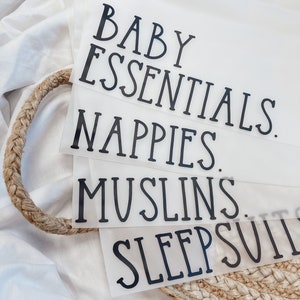 K&ouml;nnte beinhalten: Transparente Kunststoff-Organizer mit schwarzem Text, der "BABY ESSENTIALS", "NAPPIES", "MUSLINS" und "SLEEPSUITS" lautet. Die Organizer sind in einem geflochtenen Korb gestapelt, vor einem wei&szlig;en Stoffhintergrund.