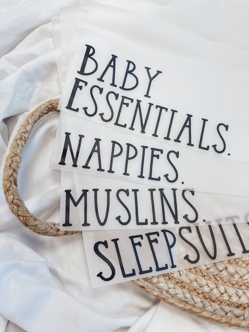 K&ouml;nnte beinhalten: Transparente Kunststoff-Organizer mit schwarzem Text, der "BABY ESSENTIALS", "NAPPIES", "MUSLINS" und "SLEEPSUITS" lautet. Die Organizer sind in einem geflochtenen Korb gestapelt, vor einem wei&szlig;en Stoffhintergrund.