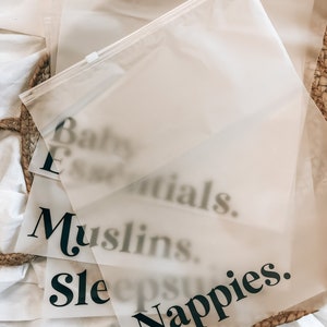 K&ouml;nnte beinhalten: Mehrere durchscheinende Plastikt&uuml;ten mit schwarzem Text, darunter "Baby Essentials", "Muslins", "Sleepsuits" und "Nappies". Die T&uuml;ten liegen auf einer wei&szlig;en Oberfl&auml;che, im Hintergrund ist ein geflochtener Korb zu sehen.