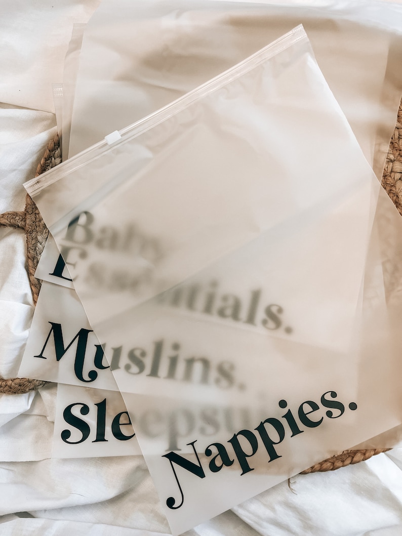 K&ouml;nnte beinhalten: Mehrere durchscheinende Plastikt&uuml;ten mit schwarzem Text, darunter "Baby Essentials", "Muslins", "Sleepsuits" und "Nappies". Die T&uuml;ten liegen auf einer wei&szlig;en Oberfl&auml;che, im Hintergrund ist ein geflochtener Korb zu sehen.