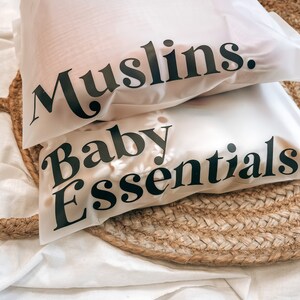 K&ouml;nnte beinhalten: Zwei wei&szlig;e, durchscheinende Beutel mit schwarzem Text, der "Muslins" und "Baby Essentials" lautet. Die Beutel liegen auf einer gewebten, kreisf&ouml;rmigen Matte und wei&szlig;em Stoff. Die Beutel scheinen Babyartikel zu enthalten.