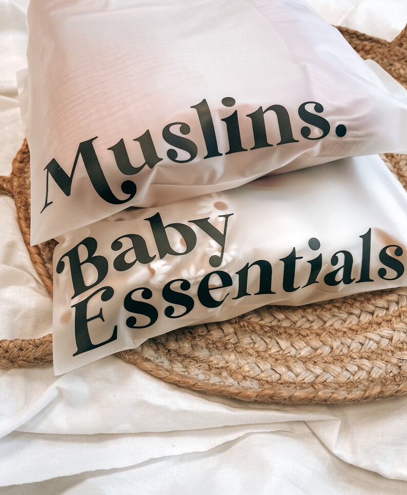 K&ouml;nnte beinhalten: Zwei wei&szlig;e, durchscheinende Beutel mit schwarzem Text, der "Muslins" und "Baby Essentials" lautet. Die Beutel liegen auf einer gewebten, kreisf&ouml;rmigen Matte und wei&szlig;em Stoff. Die Beutel scheinen Babyartikel zu enthalten.