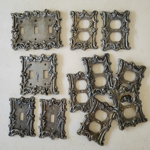 May include: A collection of antique silver-toned electrical switch plates and outlet covers. The plates feature ornate, floral, and scrollwork designs. The collection includes single and double switch plates, and double outlet covers. The plates are made of metal.