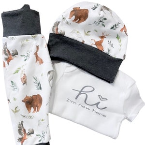 May include: A baby clothing set featuring a white onesie with the text "hi I'm new here", a matching hat, and pants. The hat and pants have a white background with a woodland animal print including bears, deer, and eagles. The cuffs are dark gray.