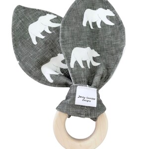 Könnte beinhalten: Grauer und weißer Hasen-Rassel mit einem Holzring. Der Stoff hat einen weißen Bären-Druck. Das Etikett lautet "Jenny Coomber Designs".