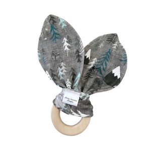 May include: A gray and teal fabric baby teether with a wooden ring. The fabric has a pattern of trees and mountains. The teether is tied with a bow and has a label that says "Jenny Chamber Designs".