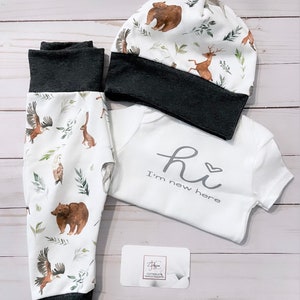 May include: A baby clothing set featuring a white onesie with the text "Hi I'm new here", matching pants, and a beanie. The pants and hat have a dark gray trim and a woodland animal print with bears, deer, and eagles.