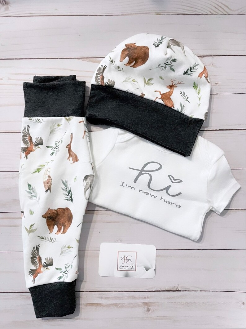 May include: A baby clothing set featuring a white onesie with the text "Hi I'm new here", matching pants, and a beanie. The pants and hat have a dark gray trim and a woodland animal print with bears, deer, and eagles.