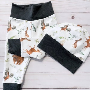 May include: A white baby pants and beanie set with a woodland animal print. The print features brown bears, deer, rabbits, and eagles with green foliage. The cuffs and waistband are dark gray.