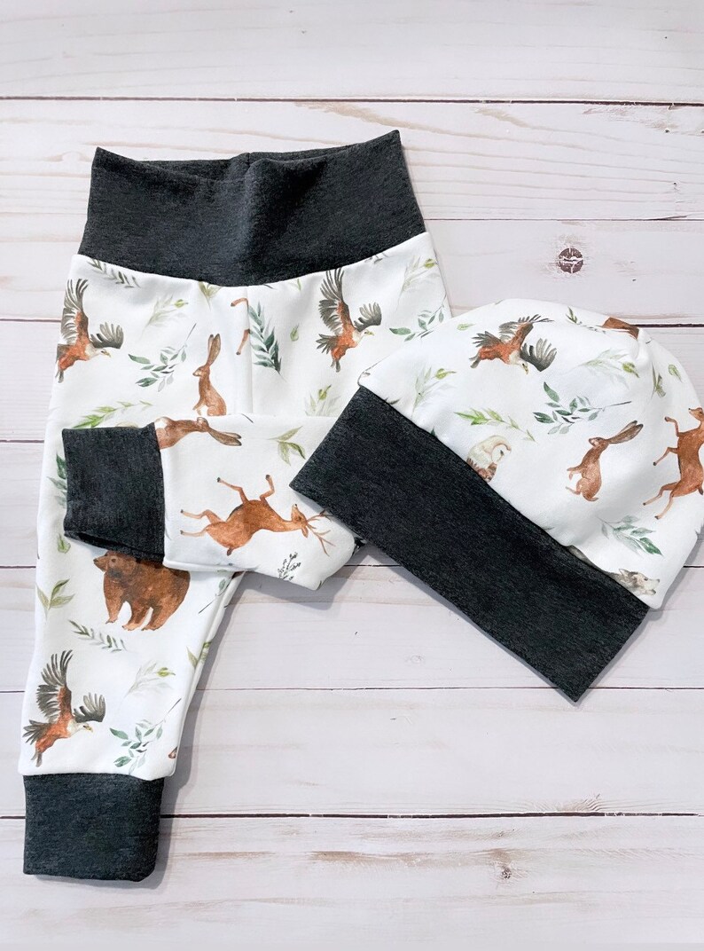 May include: A white baby pants and beanie set with a woodland animal print. The print features brown bears, deer, rabbits, and eagles with green foliage. The cuffs and waistband are dark gray.