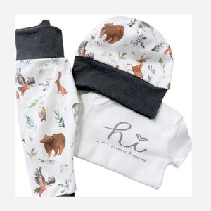 May include: A baby clothing set featuring a white onesie with the text "hi I'm new here", a matching hat, and pants. The set has a forest animal print with bears, deer, eagles, and rabbits. The cuffs and hat trim are dark gray.