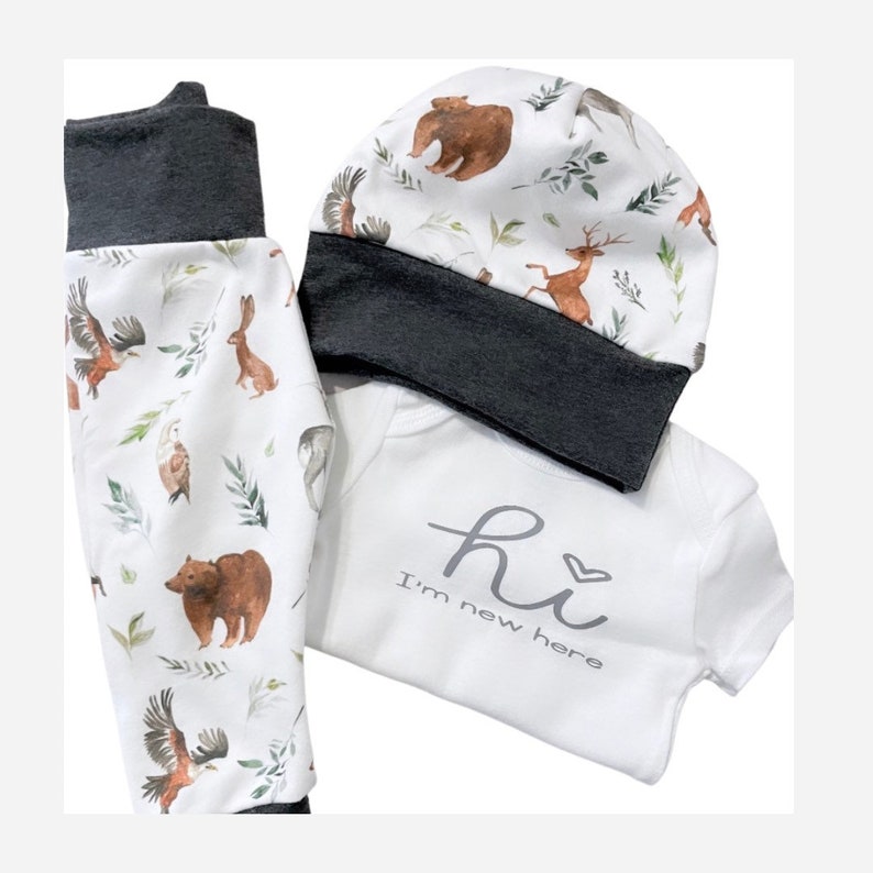 May include: A baby clothing set featuring a white onesie with the text "hi I'm new here", a matching hat, and pants. The set has a forest animal print with bears, deer, eagles, and rabbits. The cuffs and hat trim are dark gray.