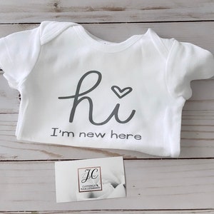 May include: White baby onesie with the text "hi" and a heart symbol, followed by "I'm new here" in gray lettering. A small business card with a logo is also visible.