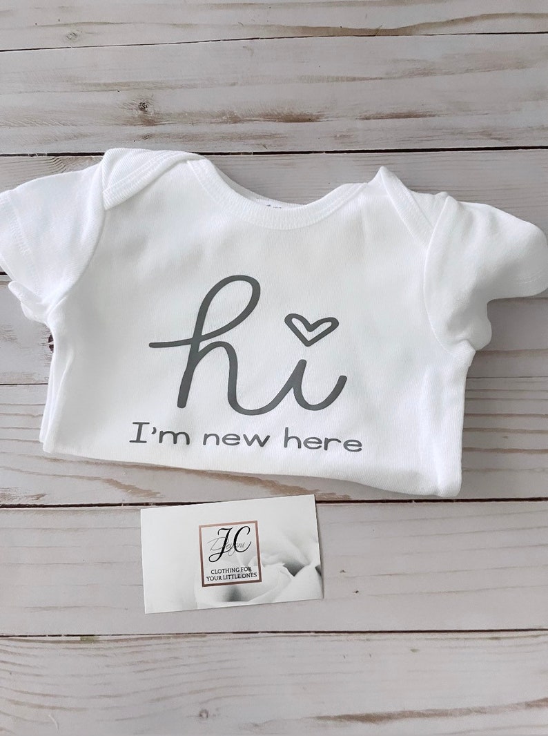 May include: White baby onesie with the text "hi" and a heart symbol, followed by "I'm new here" in gray lettering. A small business card with a logo is also visible.