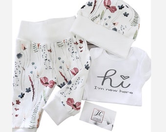 May include: A baby clothing set featuring white pants and a matching hat, both adorned with a floral pattern in shades of pink, green, and blue. A white onesie with the text "hi I'm new here" is also included.