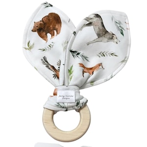 May include: A white fabric teether with a wooden ring. The fabric features a woodland animal print with a bear, wolf, fox, and eagle. The fabric is tied around the ring and has a label that reads "Jenny Coomber Designs".