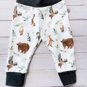 May include: White baby pants with a dark gray waistband and cuffs. The pants feature a repeating pattern of woodland animals, including bears, eagles, owls, and rabbits, with green leaf accents. The fabric is soft and comfortable.