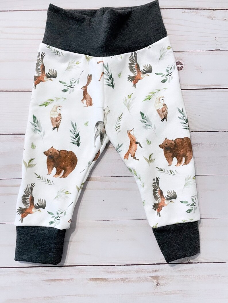 May include: White baby pants with a dark gray waistband and cuffs. The pants feature a repeating pattern of woodland animals, including bears, eagles, owls, and rabbits, with green leaf accents. The fabric is soft and comfortable.