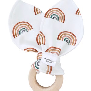 Könnte beinhalten: Ein weißer Stoff-Beißring mit Regenbogenmustern und einem Holzring. Der Stoff trägt den Aufdruck "Jenny Coomber Designs".