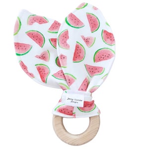 Könnte beinhalten: Ein Holz-Beißring mit einer daran befestigten weißen Stoffschlaufe. Der Stoff ist mit einem sich wiederholenden Muster aus roten und grünen Wassermelonenstücken bedruckt. Die Stoffschlaufe ist mit einem weißen Etikett versehen, auf dem "Jenny Cucumber Designs" steht.