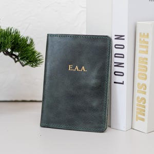 May include: A dark green leather passport holder with gold lettering that reads "E.A.A." The passport holder is next to two books, one with the word "LONDON" and the other with the words "THIS IS OUR LIFE".