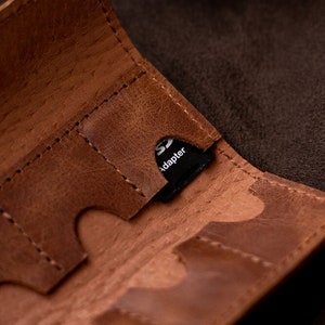 Può includere: Un porta schede SD in pelle marrone con più fessure. Un adattatore SD nero è inserito in una delle fessure. La pelle ha un aspetto invecchiato con cuciture visibili.