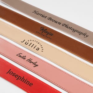 May include: Close-up of five leather camera straps in various colors: taupe, brown, beige, pink, and red. Each strap features a different name or business name in black lettering. The straps are stitched along the edges.