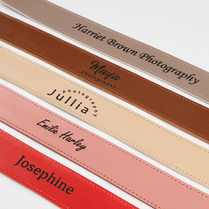 May include: Close-up of five leather camera straps in various colors: taupe, brown, beige, pink, and red. Each strap features a different name or business name in black lettering. The straps are stitched along the edges.