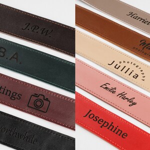 Peut inclure: Diverses lani&egrave;res de cam&eacute;ra en cuir personnalis&eacute;es, de couleurs vari&eacute;es, notamment marron, vert et bordeaux. Chaque lani&egrave;re pr&eacute;sente un texte personnalis&eacute; ou une ic&ocirc;ne d'appareil photo, avec des noms et des phrases.