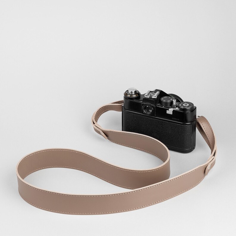 May include: A black vintage camera with a tan leather strap. The camera has silver accents and a series of dials and buttons. The strap is looped and lies in front of the camera.