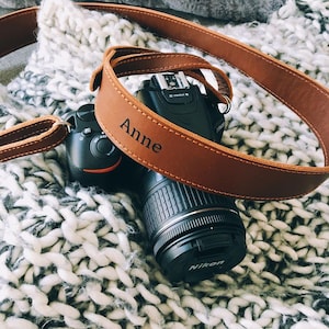 May include: A black Nikon DSLR camera with a brown leather strap. The strap is personalized with the name "Anne" and is stitched with a lighter brown thread. The camera and strap rest on a white and gray knitted blanket.