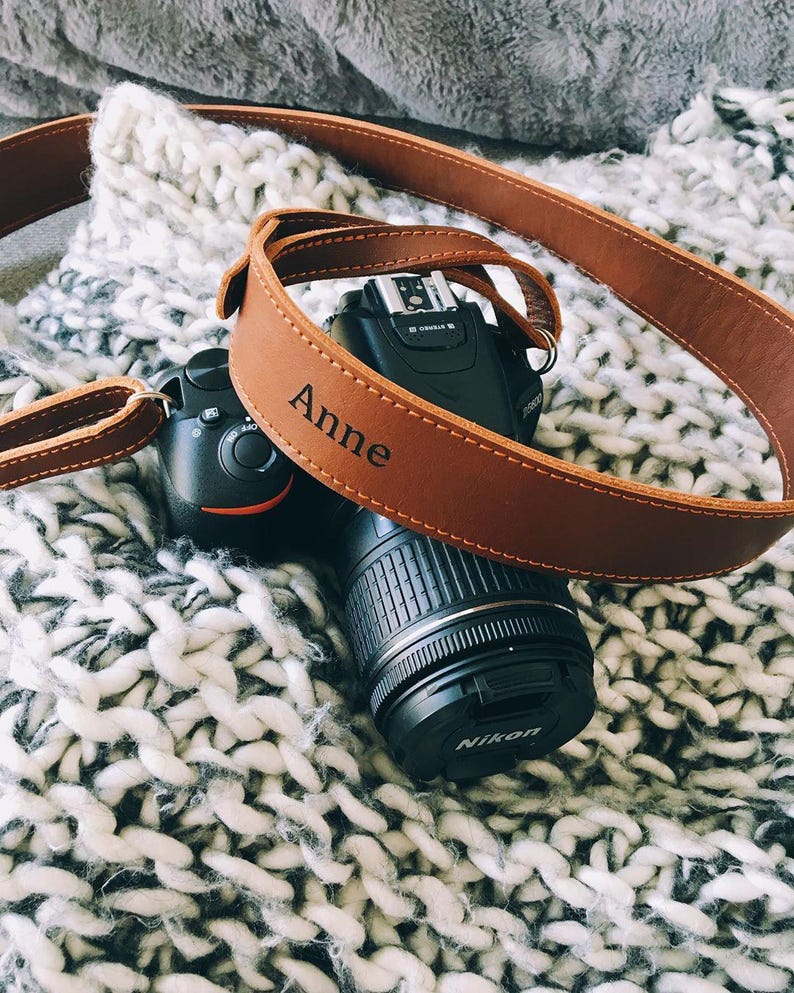 May include: A black Nikon DSLR camera with a brown leather strap. The strap is personalized with the name "Anne" and is stitched with a lighter brown thread. The camera and strap rest on a white and gray knitted blanket.